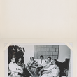 Load image into Gallery viewer, A vintage black-and-white photograph printed on a cream-colored card or page, showing several people seated in chairs around a small table in an indoor room.
