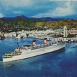 Load image into Gallery viewer, A vintage postcard-style photograph of a large white passenger liner sailing in a harbor with mountains in the background under a cloudy blue sky.
